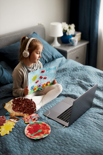 小女孩学龄前儿童学习在线显示她的作品图纸完成首页孩子学习看教训远程会说话的与导师视频调用从首页在检疫孩子使用移动PC耳机坐着床上