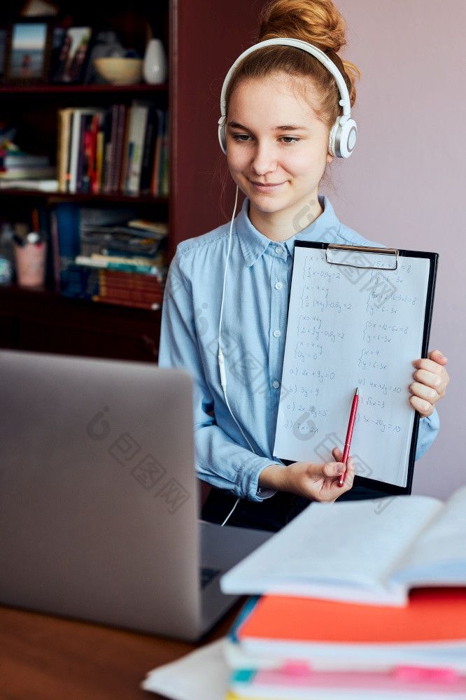 年轻的女人学生显示家庭作业有类学习在线看教训远程听教授会说话的与同学们视频调用从首页在检疫年轻的女孩使用移动PC耳机书手册坐着桌子上