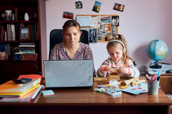 女人妈妈。工作做她的工作远程在视频闲谈，聊天调用流在线课程网络研讨会移动PC从首页而她的女儿玩与砖玩具女人坐着桌子上前面电脑看屏幕