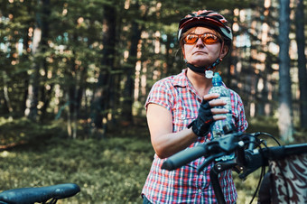 活跃的女人支出免费的夏天假期时间自行车旅行森林女人穿自行车头盔和手套持有自行车与篮子和喝水从瓶活跃的女人支出假期夏天时间自行车旅行森林