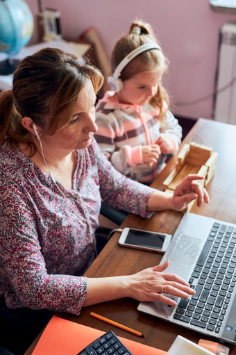 女人妈妈。工作做她的工作远程在视频闲谈，聊天调用流在线课程网络研讨会移动PC从首页而她的女儿玩与砖玩具女人坐着桌子上前面电脑看屏幕