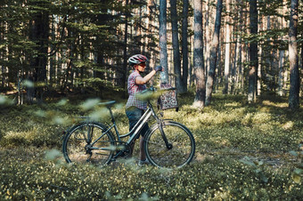 活跃的女人支出免费的假期时间自行车旅行森林阳光明媚的夏天一天女人穿自行车头盔和手套持有自行车与篮子站后面的模糊树叶子活跃的女人支出免费的夏天假期时间自行车旅行森林