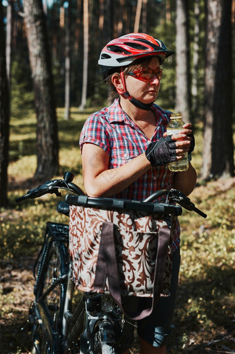 活跃的女人<strong>支出</strong>免费的夏天假期时间自行车旅行森林女人穿自行车头盔和手套持有自行车与篮子和喝水从瓶活跃的女人<strong>支出</strong>免费的夏天假期时间自行车旅行森林