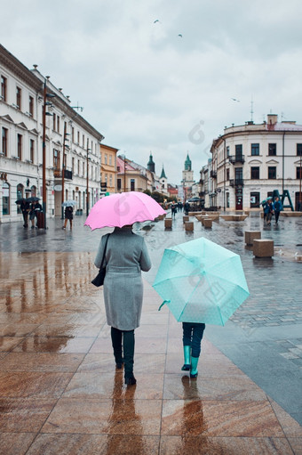 回来视图妈妈。和她的女儿持有的粉红色的和蓝色的雨伞走市中心多雨的悲观的秋天一天女人和小女孩持有的雨伞走市中心多雨的悲观的秋天一天