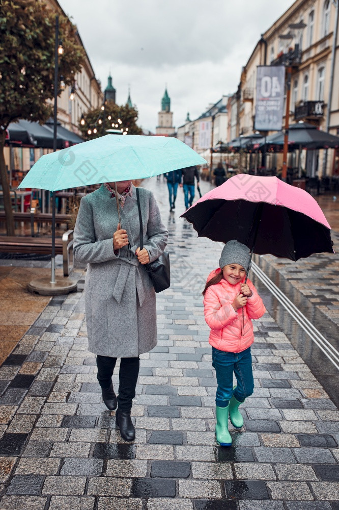 妈妈。和她的小女儿持有的粉红色的和蓝色的雨伞走市中心多雨的悲观的秋天一天女人和小女孩持有的雨伞走市中心多雨的悲观的秋天一天