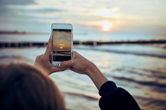 年轻的女人采取照片<strong>日</strong>落在海使用智能<strong>手机</strong>在夏天旅行后视图女孩持有智能<strong>手机</strong>和采取图片