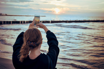 年轻的女人采取照片<strong>日</strong>落在海使用智能<strong>手机</strong>在夏天旅行后视图女孩持有智能<strong>手机</strong>和采取图片