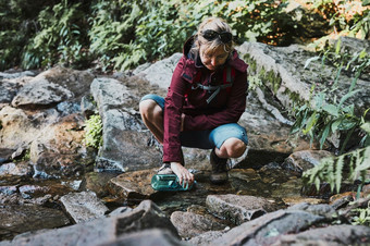 女人采取<strong>纯水</strong>瓶从山流在徒步旅行山徒步旅行者蹲岩石填充瓶与冷山水享受的在户外的夏天旅行假期