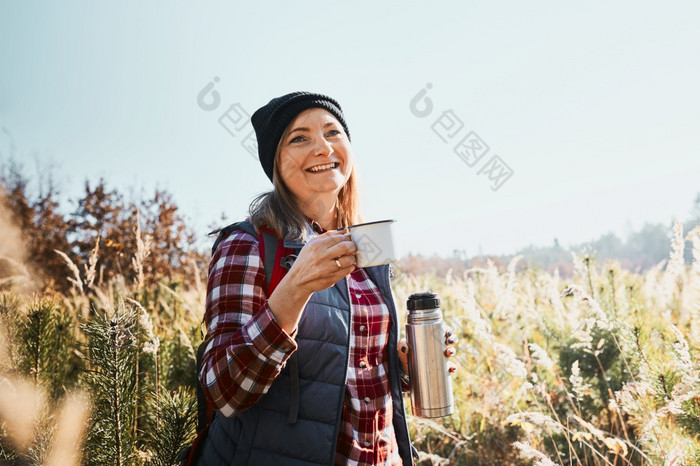 微笑女人放松和享受的咖啡在假期旅行女人站小道和看走了持有杯咖啡和热水瓶瓶女人与背包徒步旅行通过高草沿着路径山活跃的休闲时间关闭自然微笑女人放松和享受的咖啡在假期旅行女人站小道和看走了持有杯咖啡和热水瓶