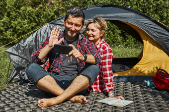 夫妇有视频调用与朋友使用智能手机而坐着帐篷野营人放松帐篷在夏天假期积极支出假期在户外关闭自然概念营生活