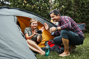 夫妇有视频调用与朋友使用智能手机而坐着帐篷野营人放松帐篷在夏天假期积极支出假期在户外关闭自然概念营生活