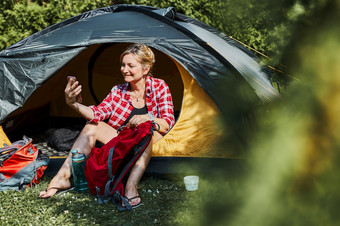 女有视频调用与朋友使用智能手机而坐着帐篷野营女人放松帐篷在夏天假期积极支出假期在户外关闭自然概念营生活