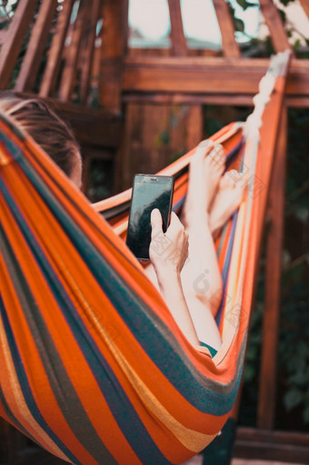 年轻的女人放松色彩斑斓的吊床和使用智能手机