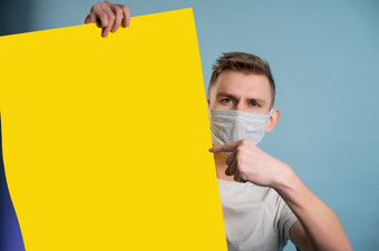 年轻的男人。穿医疗面具显示黄色的纸海报和看相机蓝色的背景广告概念年轻的男人。穿医疗面具显示黄色的纸海报和看相机蓝色的背景