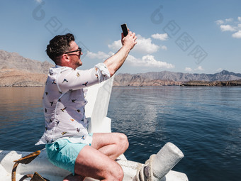 有吸引力的时尚男人。坐着船和拍摄沿海悬崖蓝色的天空和波概念休闲和旅行有吸引力的时尚男人。坐着船
