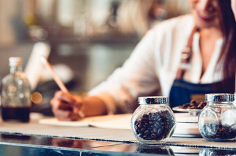 特写镜头咖啡豆瓶和快乐女咖啡师写下来报告请注意笔记本为计算业务成本和利润商店老板企业家背景咖啡商店人生活方式