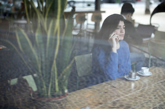 快乐女会说话的移动电话坐着咖啡馆见过通过玻璃窗口