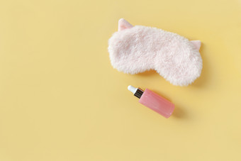 粉红色的毛茸茸的皮毛睡眠面具与小耳朵和瓶与血清为脸柔和的黄色的纸背景前视图概念生动的梦想和皮肤哪最小的风格复制空间水平