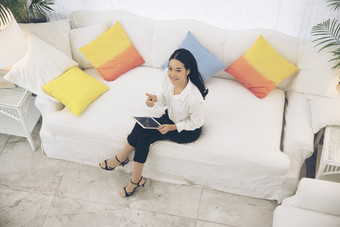 前视图年轻的美丽的亚洲女人看的相机坐着白色沙发使用平板电脑智能手机探索网站阅读文章和在线内容