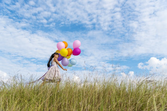 快乐的可爱的女孩持有气球运行绿色草地白色云和蓝色的天空与幸福手持有充满活力的空气气球玩生日聚会，派对快乐次夏天阳光户外