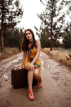 年轻的美丽的女孩黄色的衣服与色彩斑斓的围巾坐着古董手提箱浸泡泥泞的路后雨