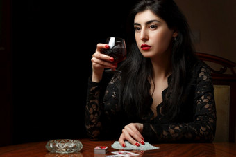 美丽的浅黑肤色的女人女人与<strong>朱红色</strong>嘴唇黑色的衣服坐着表格喝酒和玩卡片为脱衣