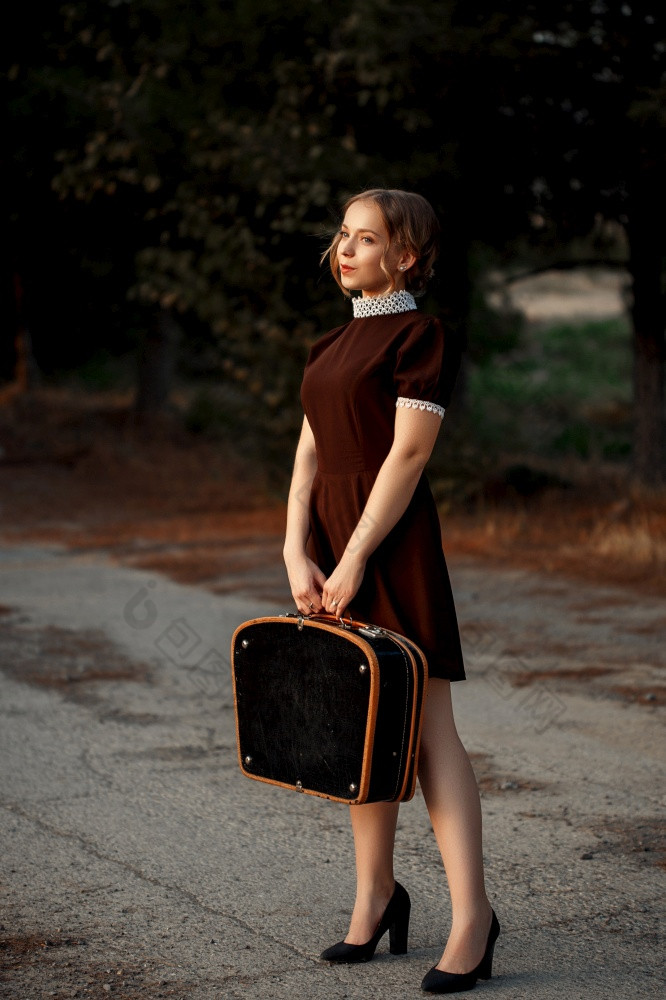 年轻的美丽的女孩棕色（的）衣服复古的风格站与黑色的手提箱她的手被遗弃的路