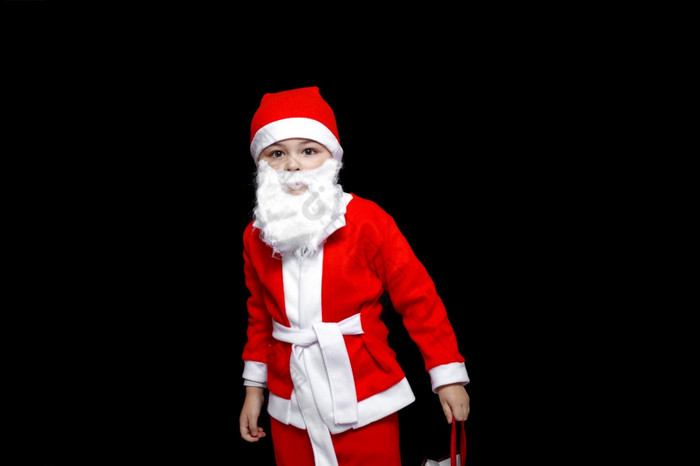 五岁的男孩有趣的圣诞老人老人服装站与包礼物黑色的孤立的背景五岁的男孩有趣的圣诞老人老人服装站与包礼物黑色的孤立的背景