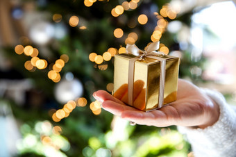 关闭手持有金礼物盒子与圣诞节树和灯的背景现在和圣诞节假期概念闪耀背景关闭手持有金礼物盒子与圣诞节树和灯的背景现在和圣诞节假期概念