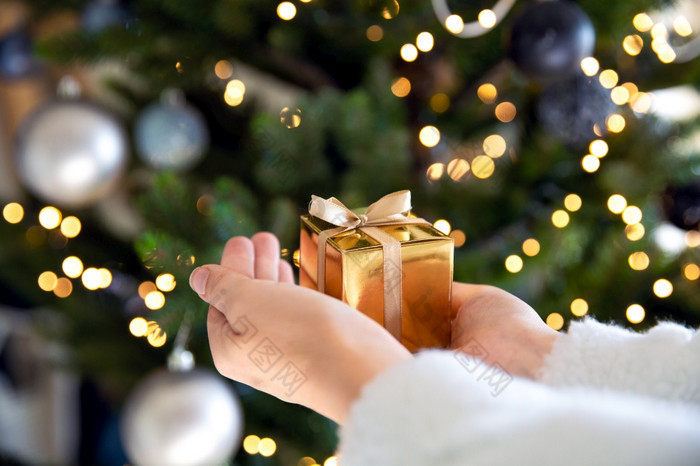 关闭手持有金礼物盒子与圣诞节树和灯的背景现在和圣诞节假期概念闪耀背景关闭手持有金礼物盒子与圣诞节树和灯的背景现在和圣诞节假期概念
