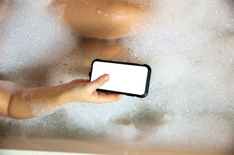 年轻的女持有空白移动电话聪明的电话浴缸与肥皂泡沫特写镜头前视图背景复制空间年轻的女持有空白移动电话聪明的电话浴缸与肥皂泡沫特写镜头前视图背景