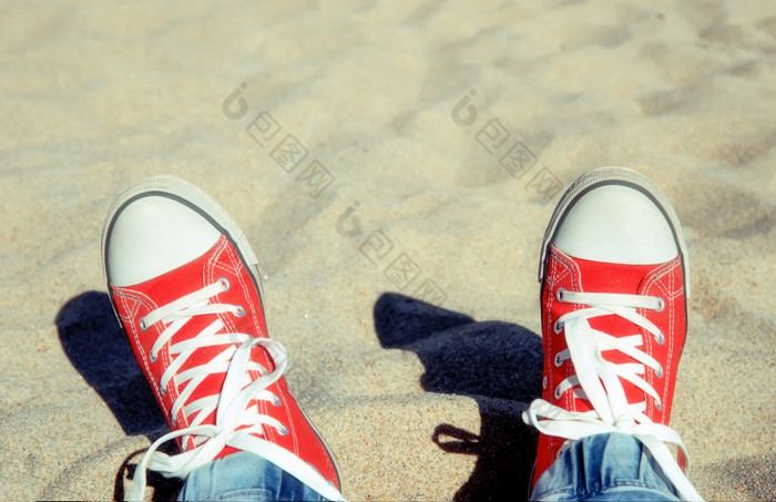 两个腿红色的运动鞋与白色鞋带对的背景光海滩沙子阳光明媚的一天两个腿红色的运动鞋与白色鞋带
