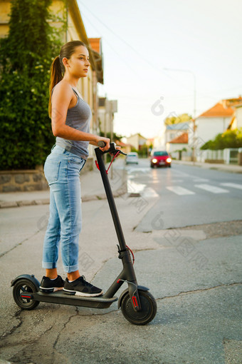 年轻的女人游乐设施电踢踏板车的街等待的十字路口穿牛仔裤夏天一天晚上交通运输