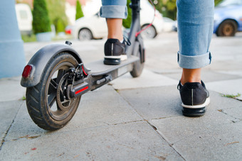 关闭女人腿脚站的电踢踏板车的人行道上穿牛仔裤和运动鞋夏天一天回来视图