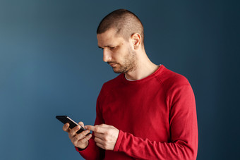 高加索人男人。穿红色的毛衣站前面的蓝色的背景墙使用聪明的电话移动发送消息短信发短信浏览互联网一边视图