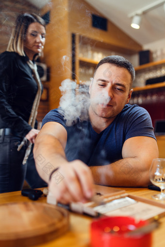 肖像年轻的高加索人男人。成人坐着表格匮乏压抑了雪茄吸烟看灰托盘穿t恤蓝色的餐厅首页而未知的女人走站后面他