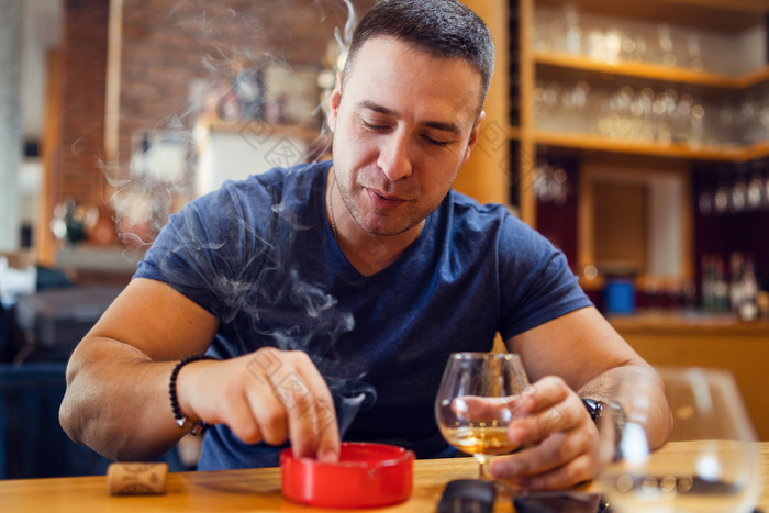 肖像年轻的高加索人男人。成人坐着表格匮乏压抑了雪茄吸烟看灰托盘穿t恤蓝色的餐厅首页持有玻璃白兰地白兰地