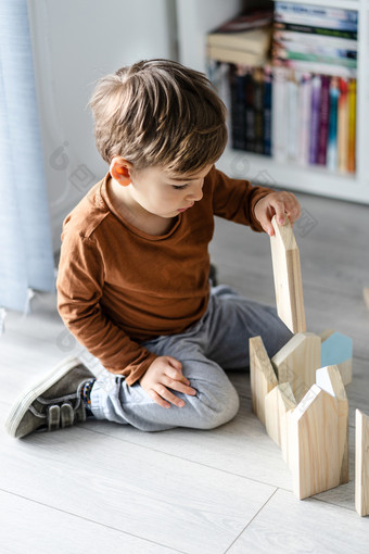 完整的长度视图小高加索人男孩小孩子首页孩子玩与木玩具砖形状房子坐着的木乙烯基层压地板上独自一人发展中创造力前视图
