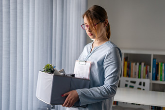 年轻的成人高加索人女人女女孩站的窗口的办公室失去她的工作持有个人项目的事情盒子被解雇了从工作驳回了由于危机经济衰退戒烟