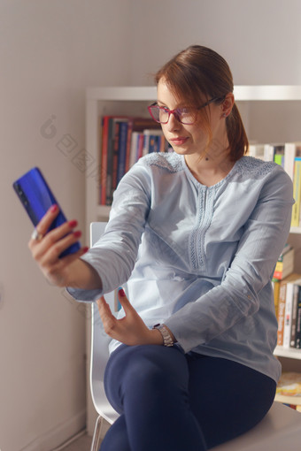 年轻的成人高加索人女人女女孩坐着的椅子首页工作前面的书架上持有智能手机移动电话采取自拍照片使视频调用一天前面视图