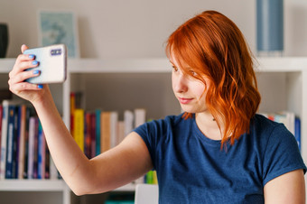 肖像美丽的年轻的女人女高加索人女孩前面的书架上橙色头发明亮的一天首页工作使用移动聪明的电话取自拍照片穿蓝色的t恤