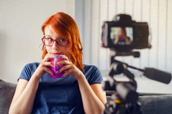 年轻的女高加索人女人博主坐着前面相机首页办公室工作视频博客喝杯茶咖啡穿t恤和眼镜看的相机前面视图