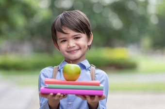 可爱的小男孩穿蓝色的衬衫和携带学生背包收到苹果从妈妈。为早餐之前学校早餐重要的为孩子发展健康和健康