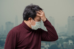 亚洲男人。穿的脸面具对空气污染与手捕捉的头疼的阳台高公寓哪一个可以看到污染和重雾在的曼谷城市景观背景