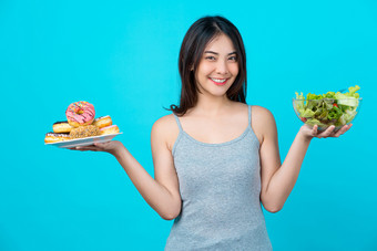 有吸引力的亚洲年轻的女人持有和选择之间的磁盘甜甜圈蔬菜沙拉眼镜碗孤立的蓝色的颜色背景重量损失和<strong>避免</strong>垃圾食物为节食和健康的