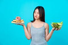有吸引力的亚洲年轻的女人持有和选择之间的磁盘甜甜圈蔬菜沙拉眼镜碗孤立的蓝色的颜色背景重量损失和避免垃圾食物为节食和健康的
