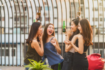 幸福亚洲女孩朋友集团庆祝和跳舞在一起与她的啤酒瓶日落时间屋顶市中心酒店夜总会母鸡晚上假期每年周年纪念日聚会，派对概念