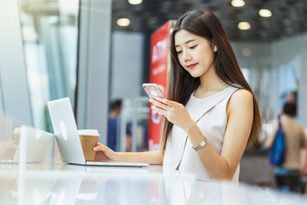 亚洲年轻的女持有杯咖啡和使用移动电话与技术移动PC现代咖啡商店共同办公空间技术聪明的移动和赶时髦的人生活方式企业家概念