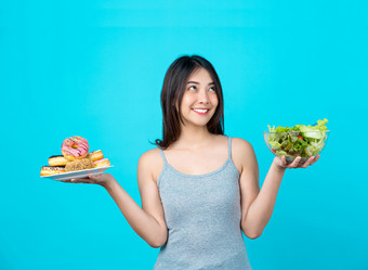 有吸引力的亚洲年轻的女人持有和选择之间的磁盘甜甜圈蔬菜沙拉眼镜碗孤立的蓝色的颜色背景重量损失和<strong>避免</strong>垃圾食物为节食和健康的