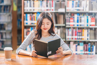 亚洲年轻的学生休闲西装阅读的书与杯咖啡图书馆大学通知书的的木表格在的书架子上背景回来学校概念